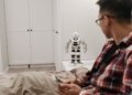 A man examines a robotic toy placed on a table in a contemporary bedroom setting.