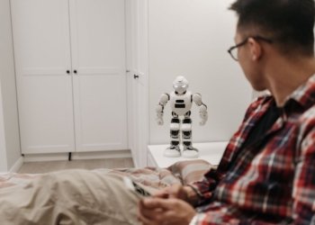 A man examines a robotic toy placed on a table in a contemporary bedroom setting.