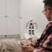 A man examines a robotic toy placed on a table in a contemporary bedroom setting.